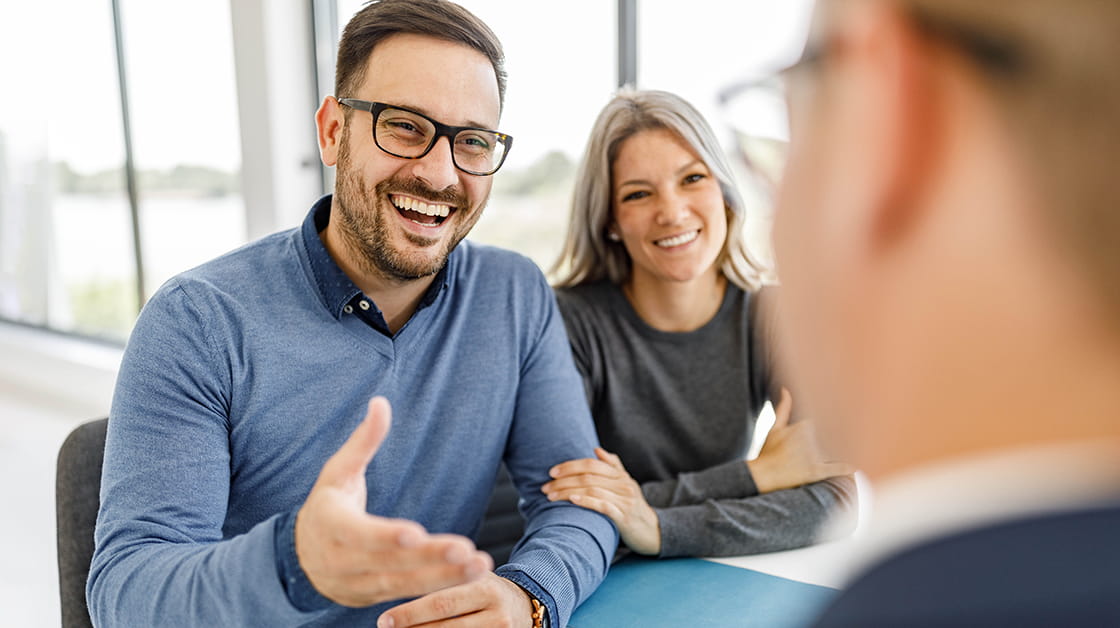 Two smiling clients sit across a desk from their solicitor. On the left sits a white man with short dark brown hair, a beard and glasses, wearing a blue jumper over a blue shirt. Next to him sits a white woman with long, wavy silver hair wearing a grey shirt. Their solicitor's profile is blurred and faces away from the camera. 