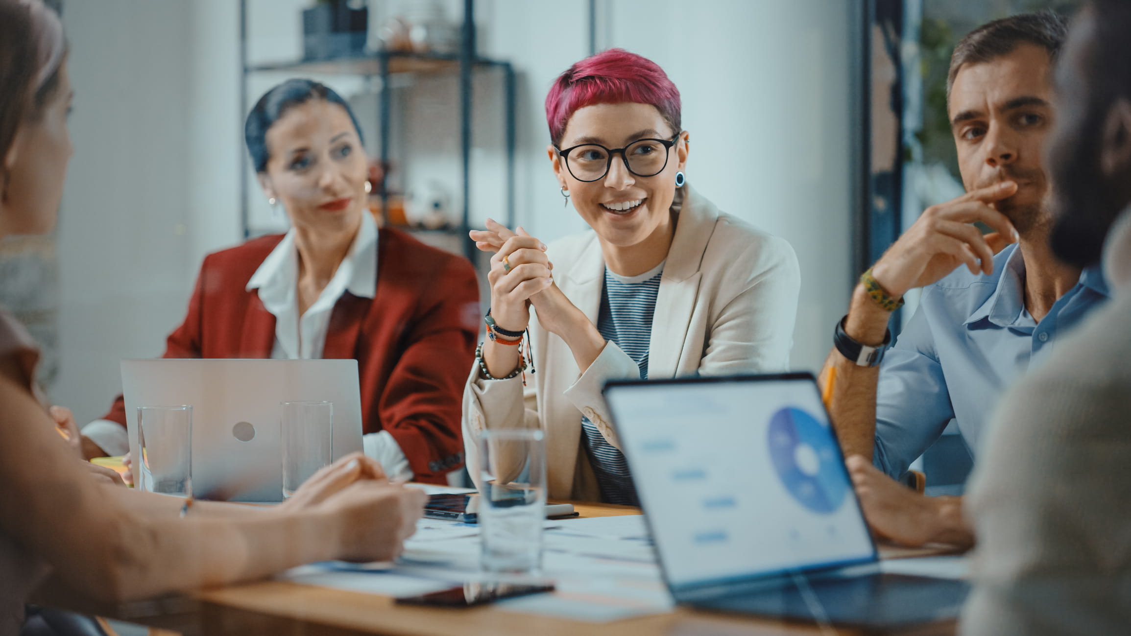 A woman with glasses and pink hair sits at a boardroom table with colleagues.