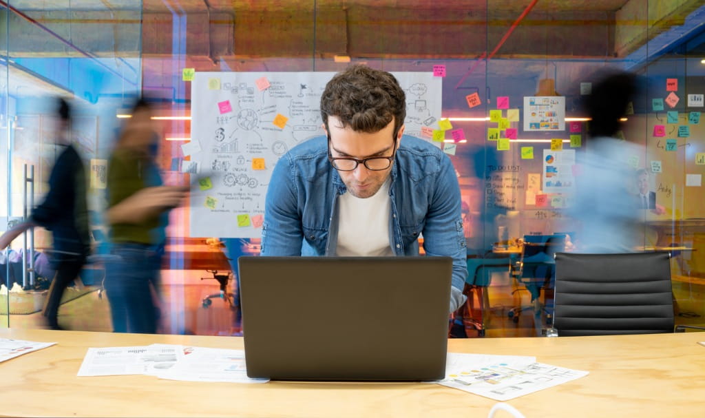 Man working at a creative office using his computer and people moving at the background 