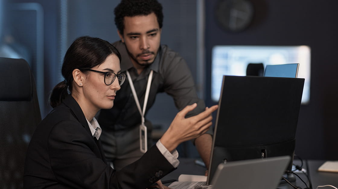 A woman with brown hair and glasses is sitting at a desk looking at a computer, a man is behind her, leaning on the desk.