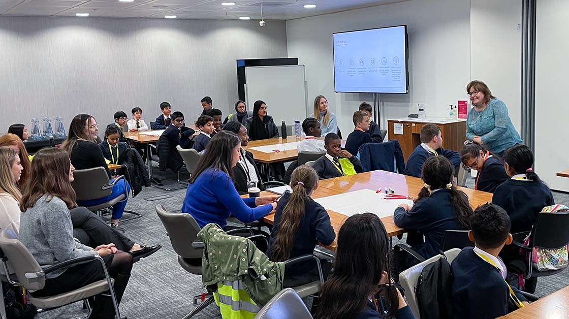 Students and volunteers in a classroom sitting around small group tables listening to a teacher.