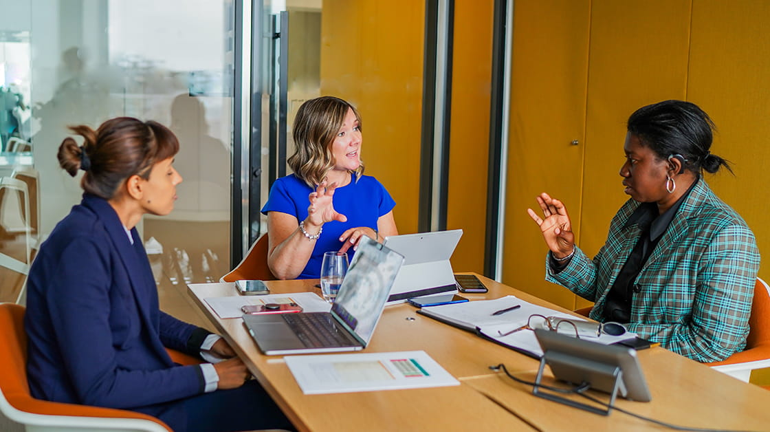 Three colleagues speaking in a meeting room. The person on the left is sitting with a laptop in front of them, wearing a navy blue suit with short brown hair tied up. In the middle, a person with short brown hair, wearing a blue dress is gesticulating in front of them, they have a laptop on the desk in front of them. On the right is a person with short black hair and a hearing aid. They are wearing a black blouse and green checked jacket, They are gesticulating in front of them.