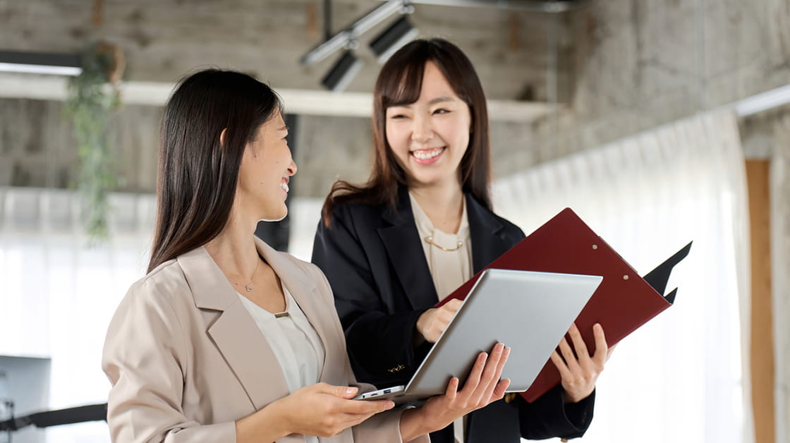 Two Asian business women, with long brown hair and standing in an office and smiling. The woman in the foreground is wearing a beige jacket and white blouse, the other woman smiling at her is wearing a black jacket and holding a folder.