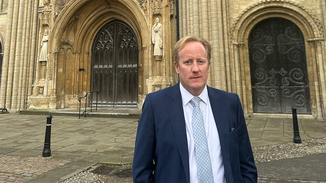 Dan Chapman stands in front of Norwich Cathedral. Dan is a white man with short, blonde, wavy hair. He wears a dark navy suit, white shirt and silver tie.