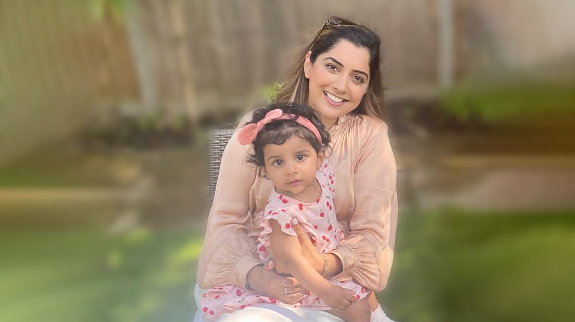 Amandeep Khasriya, a woman of South Asian heritage with long dark brown hair, sits on a garden chair with her young daughter on her lap. She is smiling widely and wears a peach blouse, white trousers and brown sunglasses on the top of her head. Her daughter wears a pink patterned dress and a pink bow on her head.