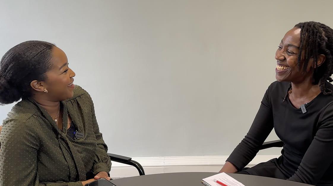Mary and Angela are two black women. They are sitting at a table conversing. Mary is wearing a green blouse and a silver necklace. She has her hair tied back. On her right, Angela is wearing a black dress. She has her hair down. They are both smiling at each other. 
