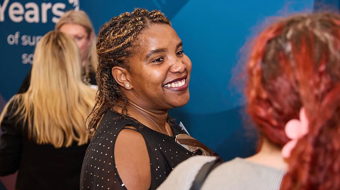 A Black woman with braids is smiling at an event, chatting with other people.