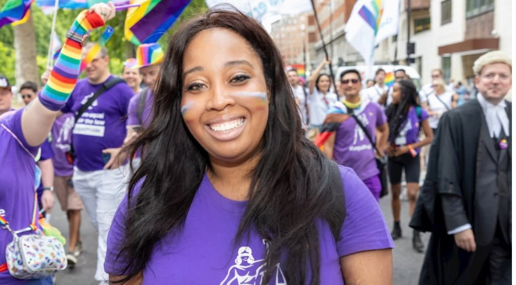 Jacqui is a Black woman with long black hair. She is standing in front of a crowd wearing a purple t-shirt and face paint. 