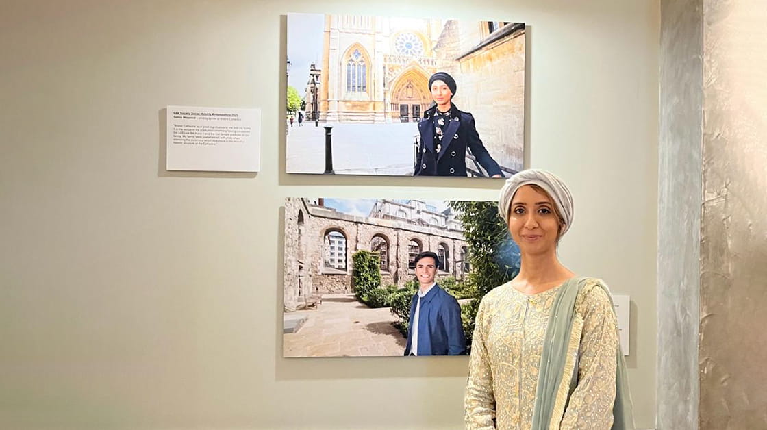 Salma Maqsood stands smiling next to her portrait at the Law Society in Chancery Lane, London. Salma is a woman of South Asian heritage wearing an ornately decorated gold and sage long-sleeved dress, a sage scarf over her shoulder and a beige hijab.