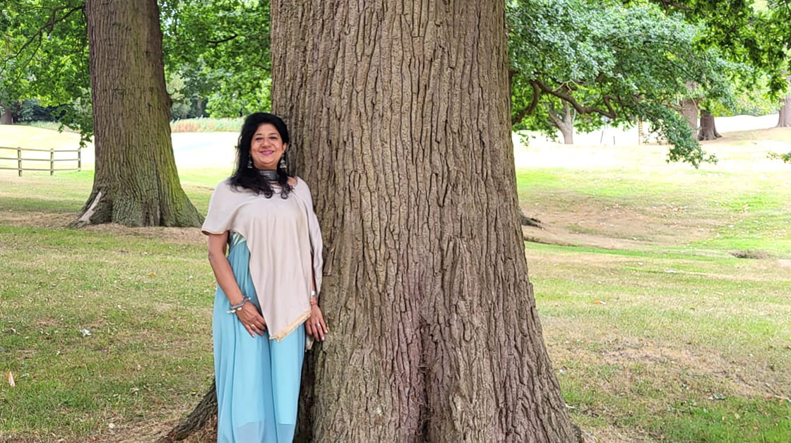 Shama Gupta is a woman of Indian heritage with long straight black hair. She is smiling and wears a long pale bright blue dress and beige shawl. She stands in parkland next to a large tree.