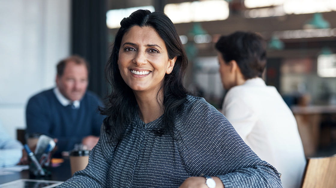 A smiling woman of South Asian heritage sits in a meeting room with two other colleagues. She has long wavy dark brown hair and wears a patterned navy blouse and silver watch.