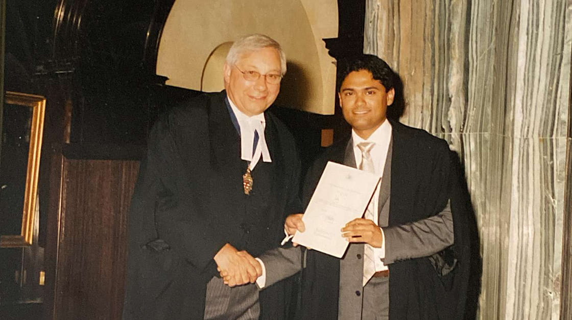 Susanta Banerjee (right) shakes hands with a representative of the Law Society at his admission ceremony. Susanta is a man of South Asian heritage with short black hair. He is smiling and wears a black graduation robes over a grey suit, white shirt and silver tie.