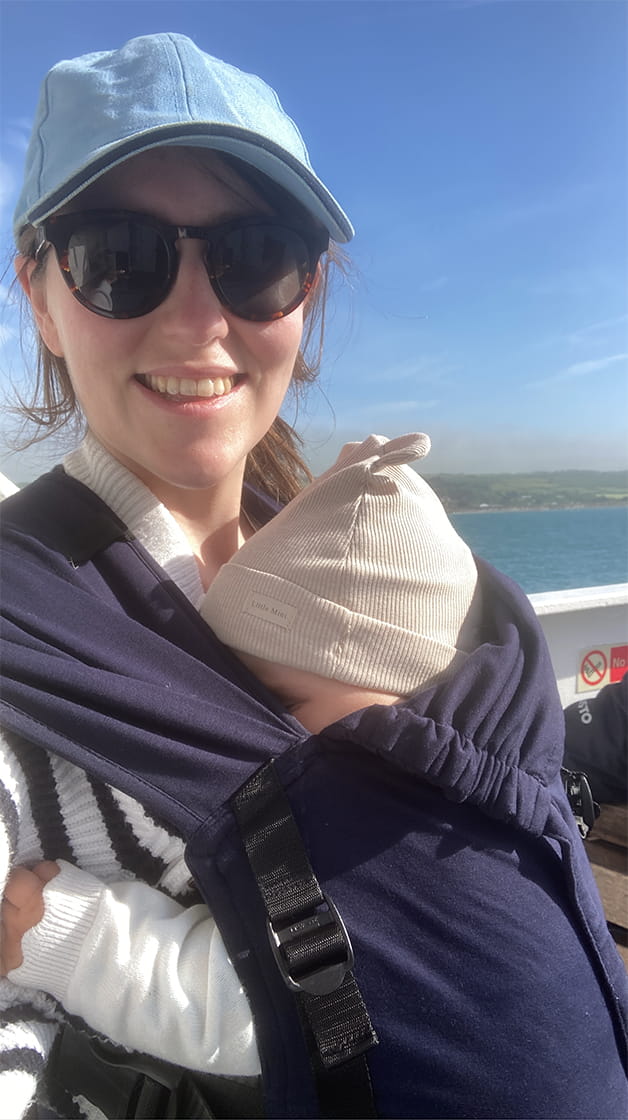 Anna Vroobel is a white woman with long, dark brown hair tied up in a ponytail. She wears a blue and white striped sweatshirt, a blue basketball cap and dark sunglasses, carrying a small baby in a navy sling.