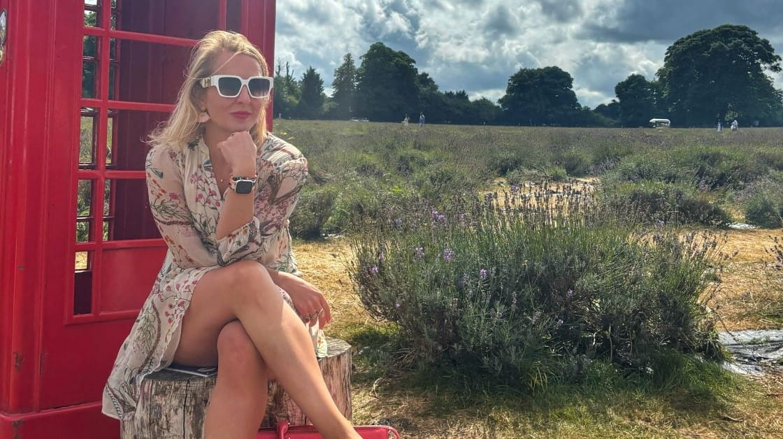 Kimberley Whitaker is a white woman with shoulder length straight blonde hair. She sits outside in front of a red telephone box, wearing a floral patterned dress and white sunglasses.