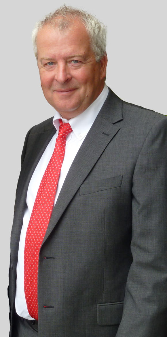 Nigel is an older white man, with white hair. He is wearing a suit with a red tie.