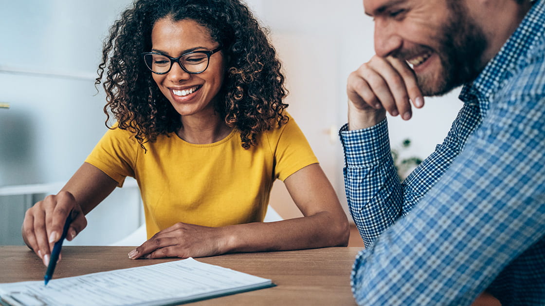 A smiling Black woman with long, curly, dark brown hair sits with a smiling white man with short brown hair and a cropped beard. She wears glasses and a yellow shirt. He wears a blue checked shirt.
