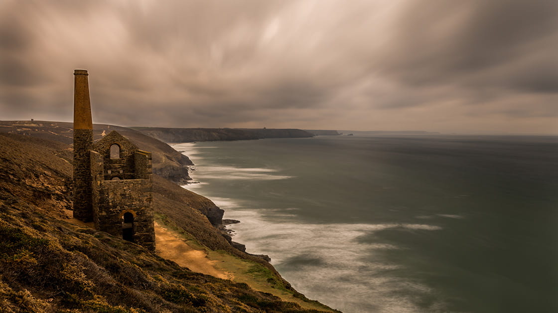 An old building sits atop a cliff on the left of the image. To the right, the coastline and a choppy seascape. The sky is dark and moody. 