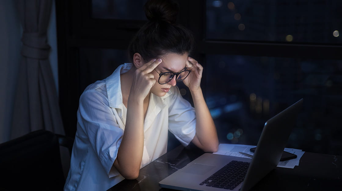 Young woman working at laptop late at night, rubbing her temples with her eyes closed
