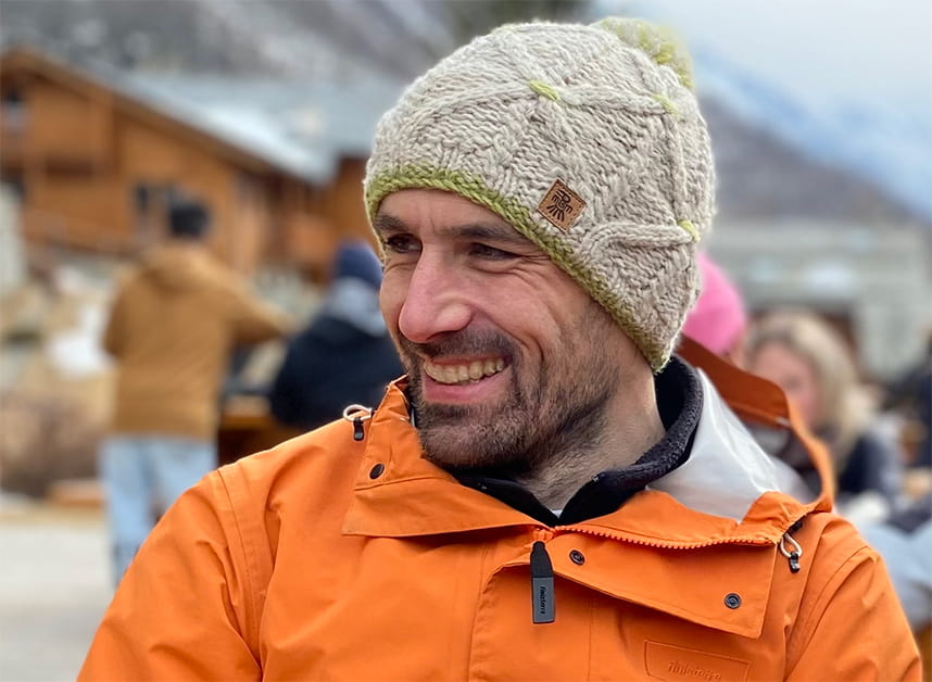Tim Pritchard is a white man with a dark beard wearing a beige bobble hat and an orange winter jacket. He sits in a seating area high in the mountains, surrounded by people.