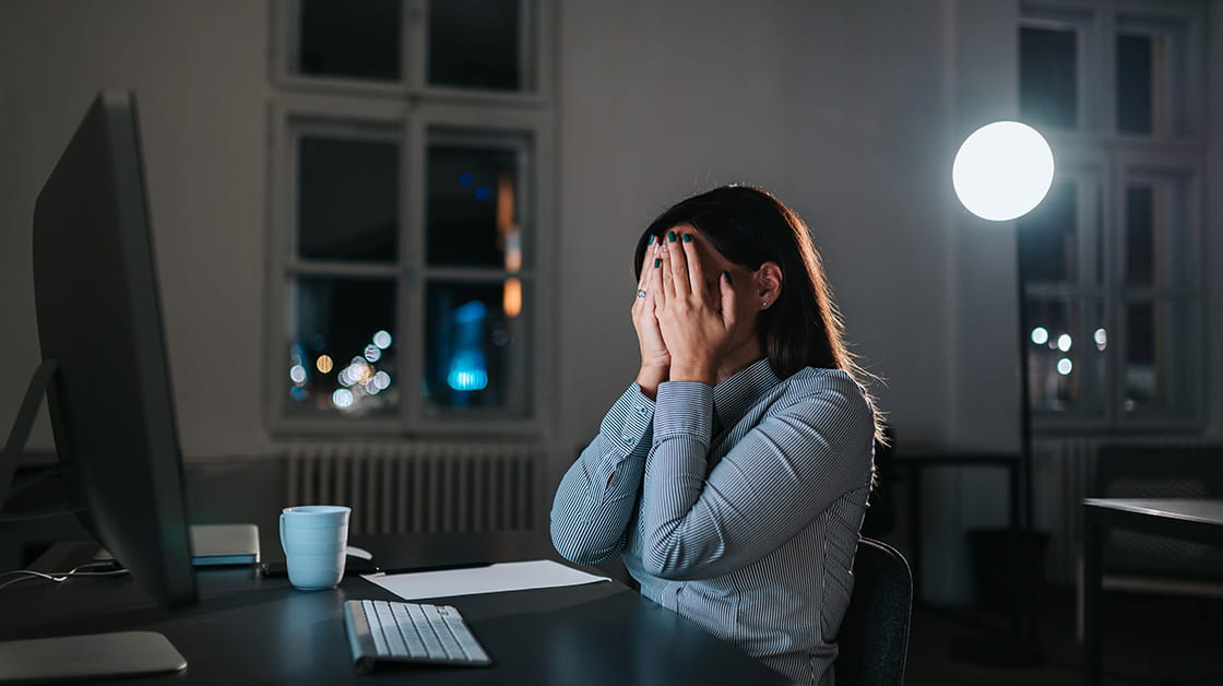 A woman sits in a darkened office and covers her face with her hands.