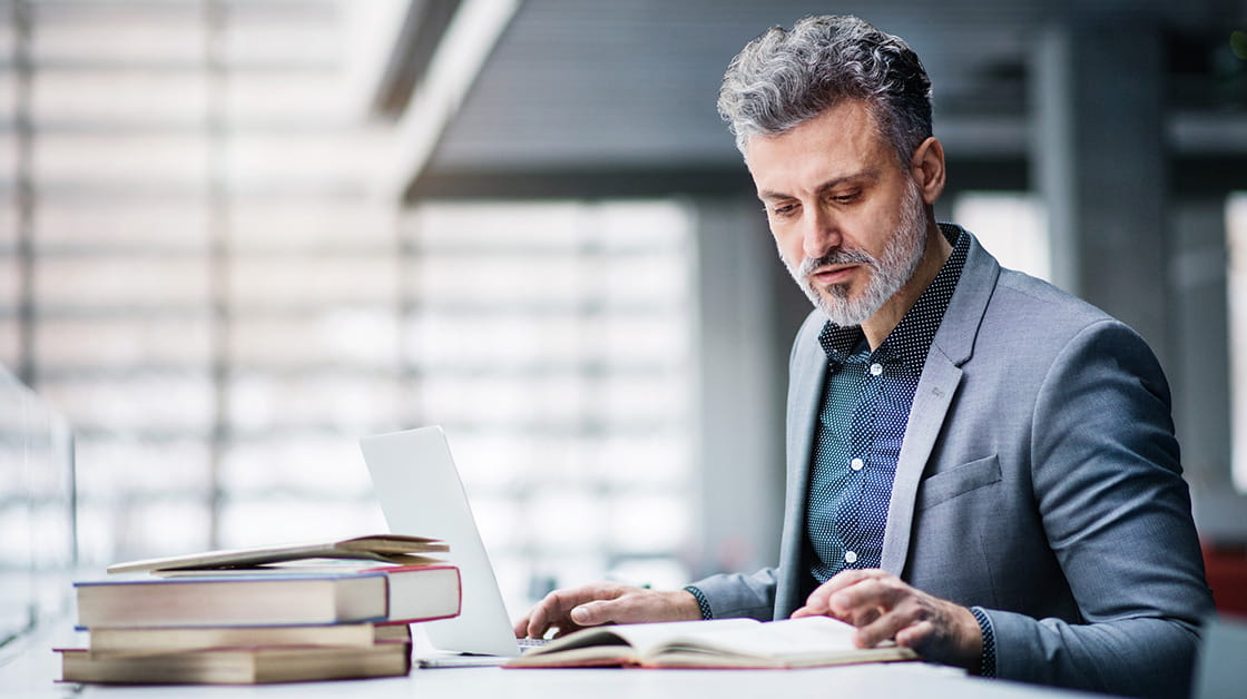 A man with grey hair and a beard sits at a desk reviewing a textbook.