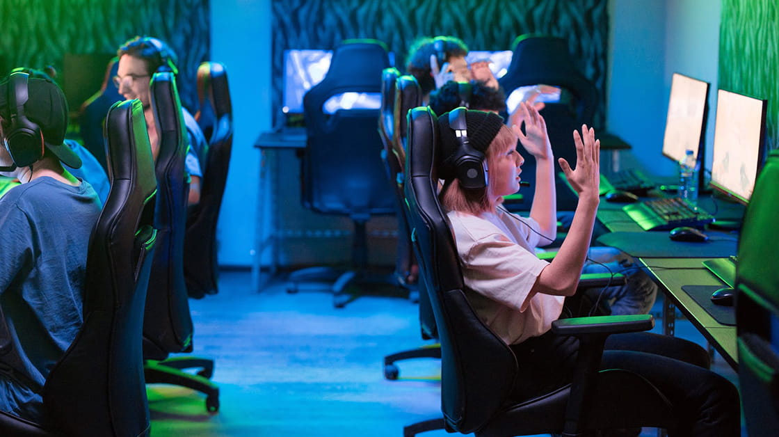 Young people sit at two rows of gaming computers - they wear headsets, the chairs have headrests and the room is lit by green neon light