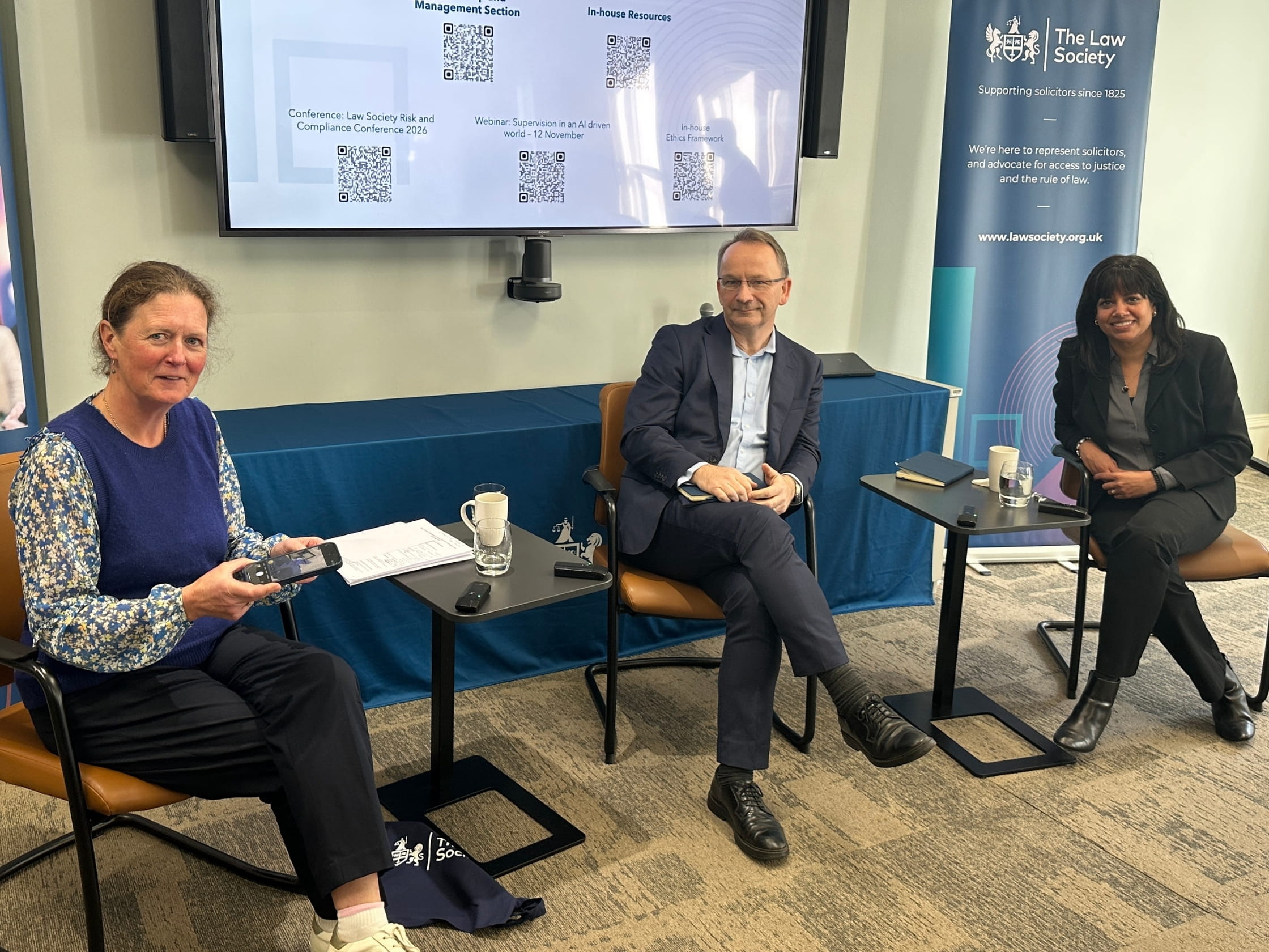 Three people sit in seats, in front of a large screen and Law Society banner.
