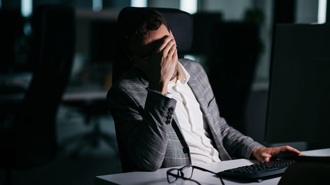 A man sits in a dark office, in front of a PC, rubbing his eyes, as if stressed.