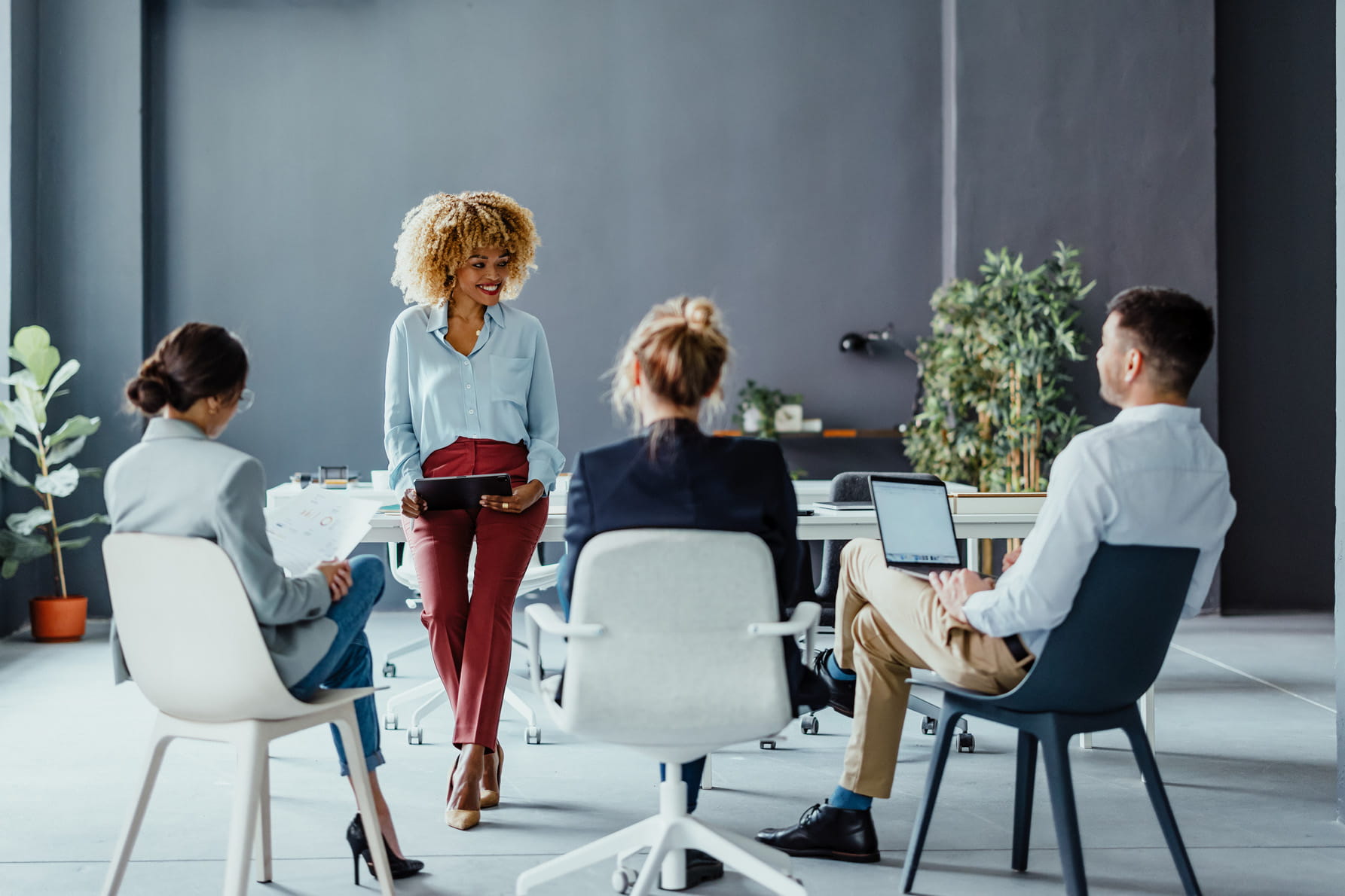A woman leans against a boardroom table and speaks to three colleagues who are all sitting on chairs.