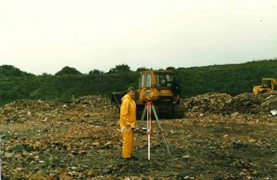 Joe Kelly wears yellow overalls and stands on a construction site in front of a JCB