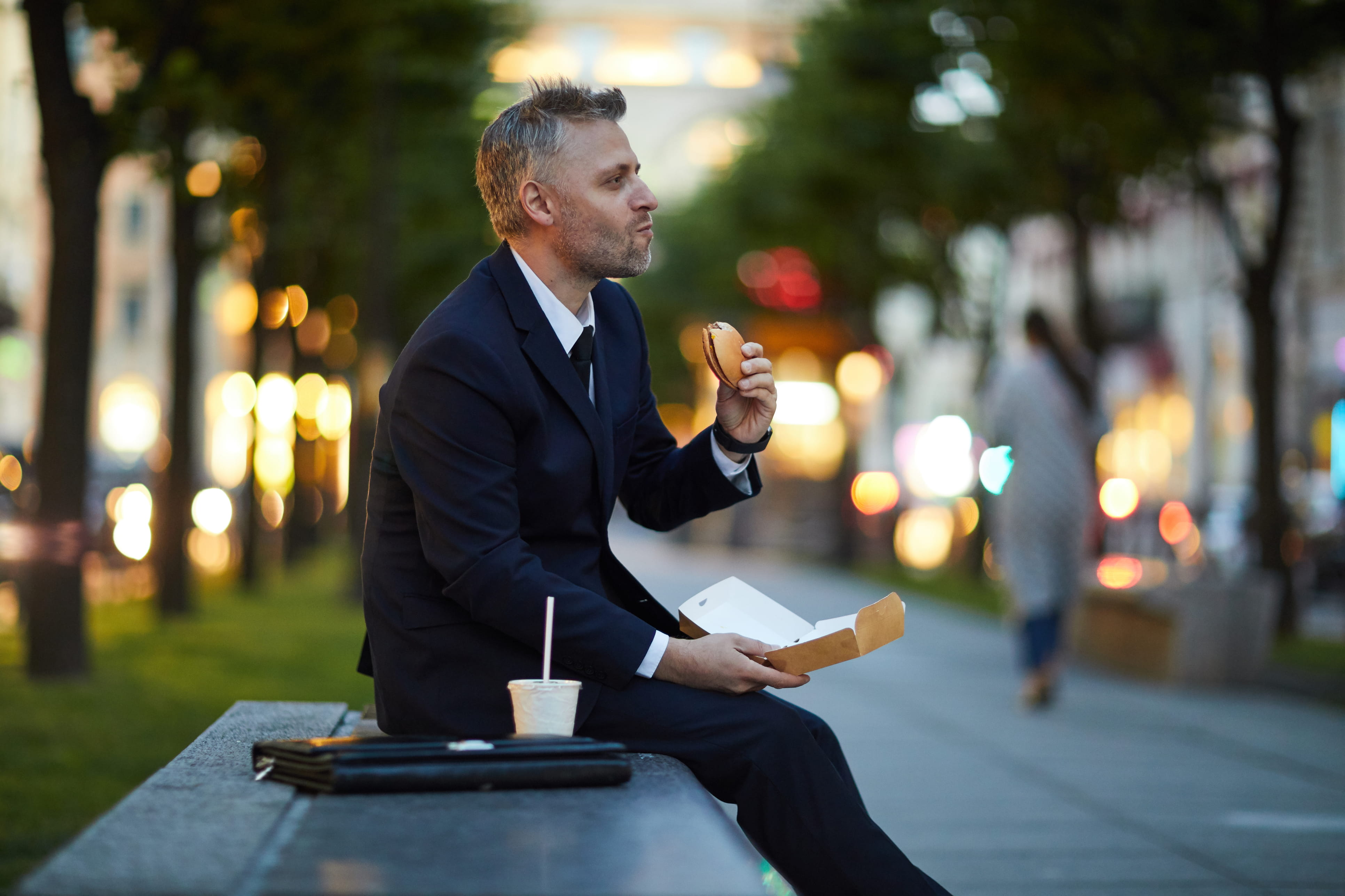 A white man in a suit sits on a park bench eating a burger. 