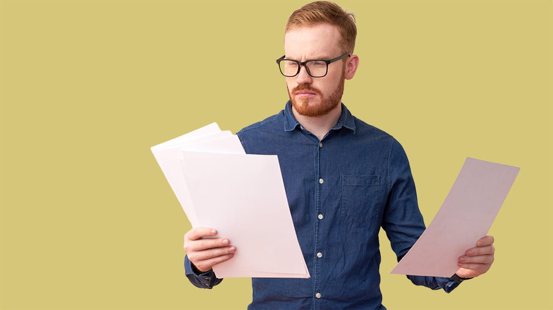 A man with red hair and glasses looks at documents in each hand. 