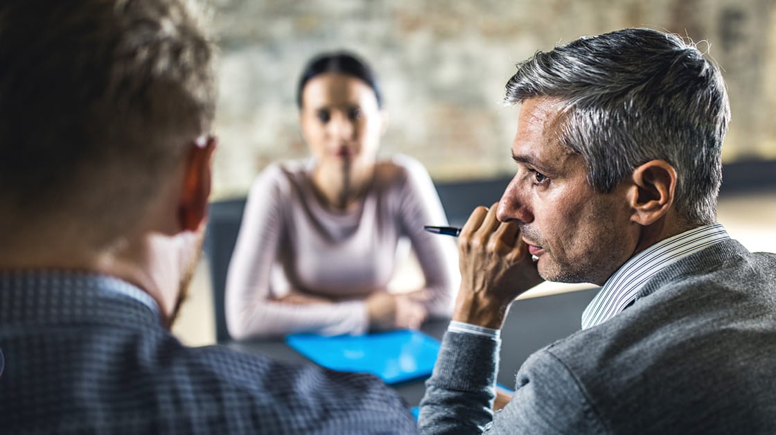 A man with grey hair whispers to another man at a board table.