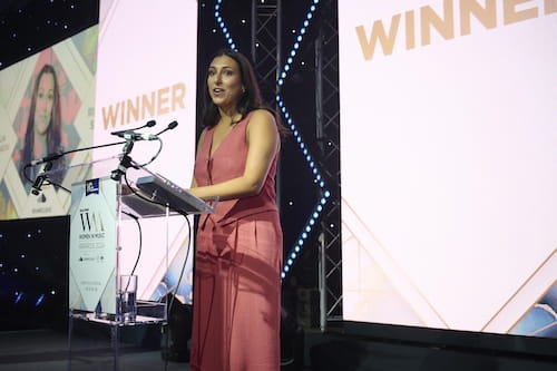 Talia Shabatai giving a speech, in front of signs that say 'winner'.