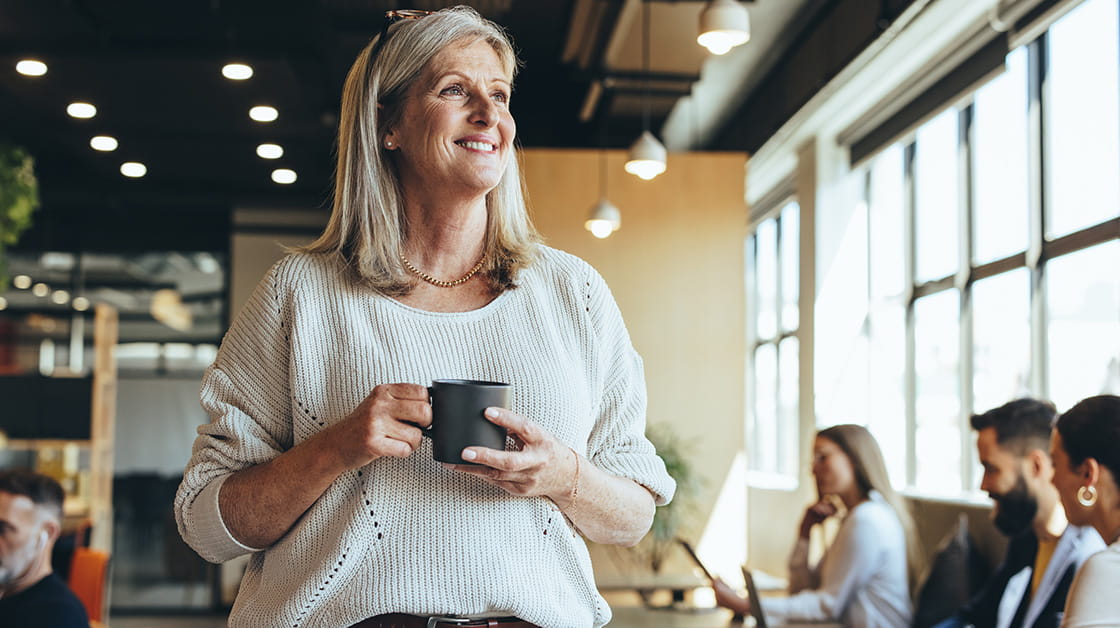 A woman with blonde hair stands next to a bank of desks in an office, she is smiling and holiding a mug.