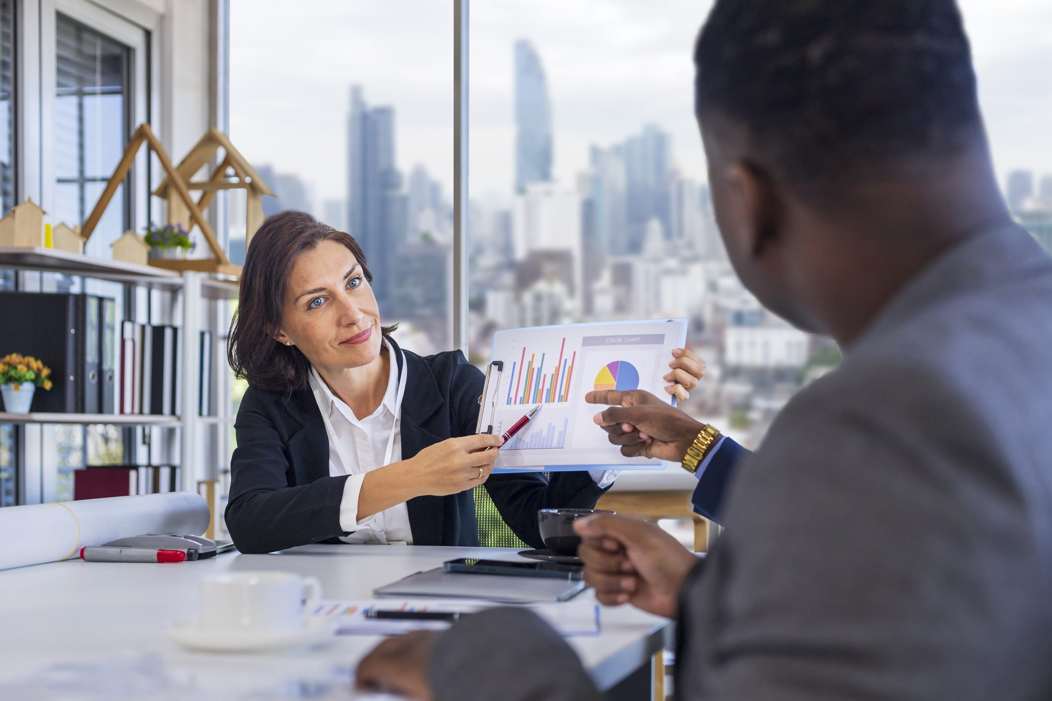 Woman manager is showing annual report chart to her African American colleagues in the executive meeting for next year plan with city skyline background for global business and investment