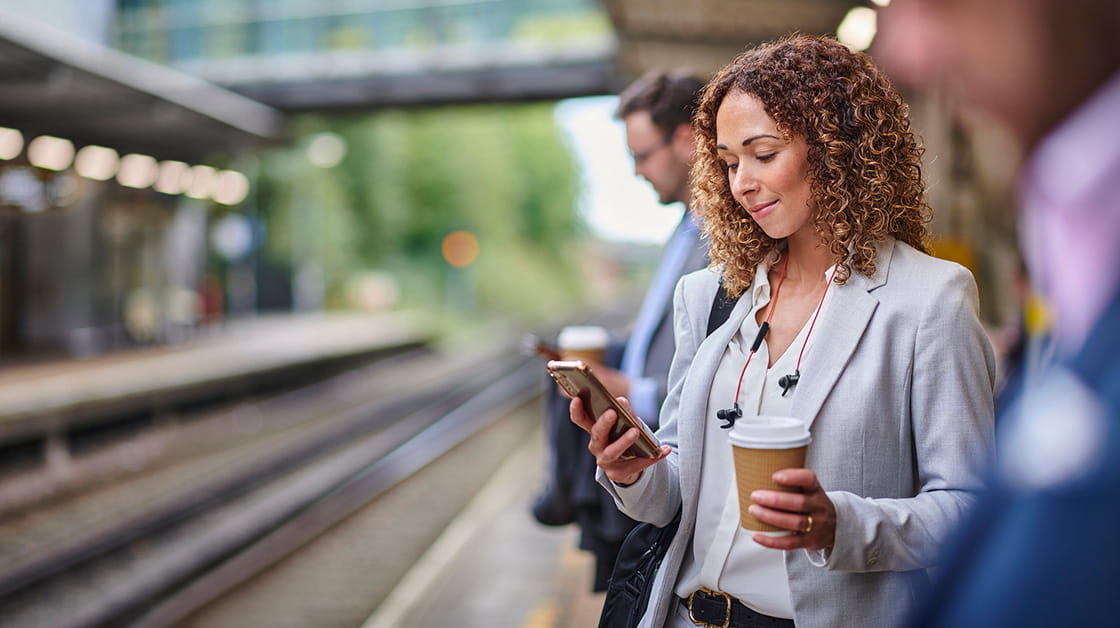 a woman dressed for work, stands on a train platform, with her one in one hand and a takeaway coffee in another. 