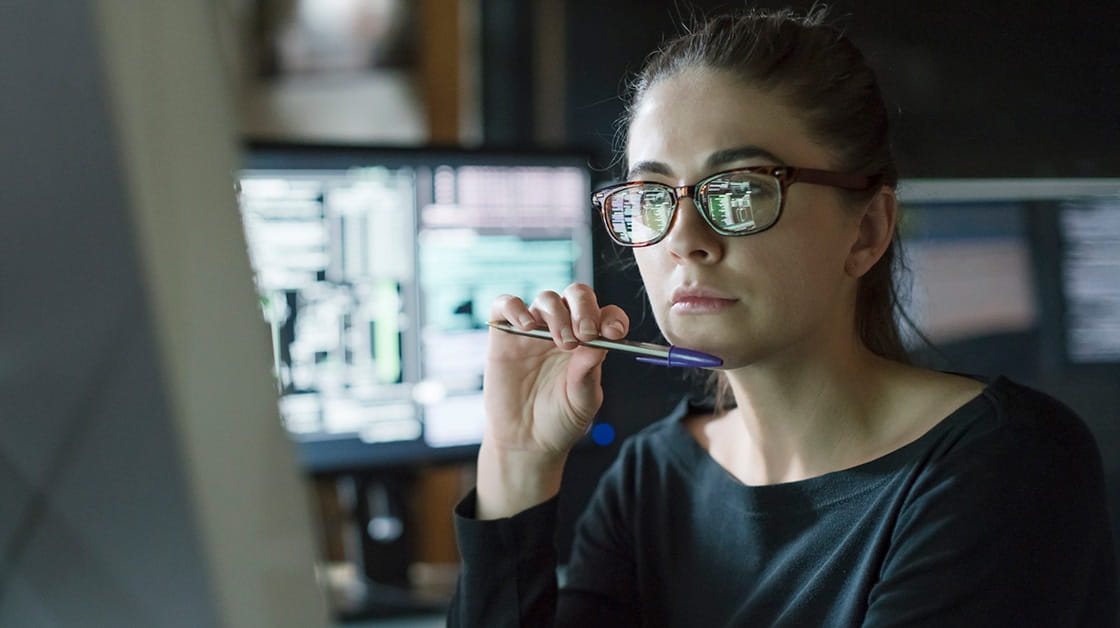 A woman with brown hair and glasses looks at computer screen in a dark office.