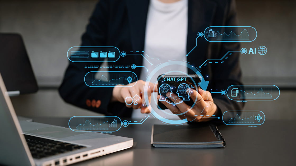 A woman sits in front of a laptop holding a mobile phone