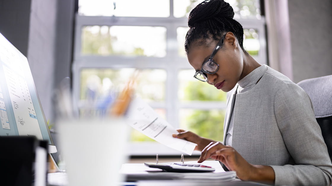 A woman sits at a desk infront of her computer and works on a calculator.
