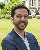 Marco Cillario is a white man with brown hair. He is standing outside wearing a navy blue suit, with his arms crossed and smiling.