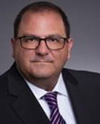 Michael Padua is a white man with closely cropped dark brown hair. He wears glasses, a black suit, a white shirt and a red striped tie.