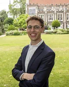 Will Miller is a white man with short straight brown hair. He stands in Lincolns Inn, smiling widely and wearing a navy suit and open-collar white shirt.