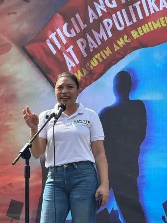 Human rights lawyer Kristina Conti stands on stage and talks into a microphone. She is wearing blue jeans and a white t-shirt and is speaking passionately
