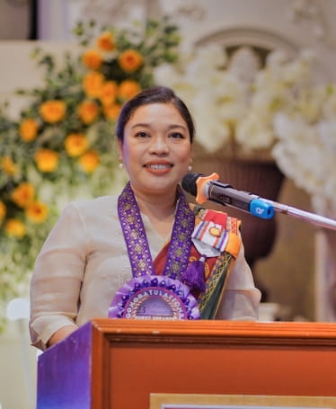 Human rights lawyer Kristina Conti is dressed in finery and purple ribbons and rosettes. She is standing at a lectern in front of a microphone and delivering a speech