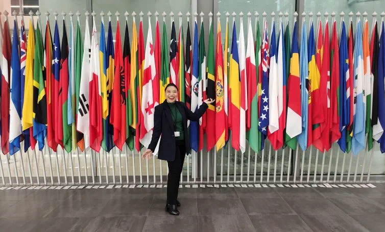 Human rights lawyer stands in front of a collection of flags at the International Criminal Court. She is wearing a black suit with a green top and is smiling.