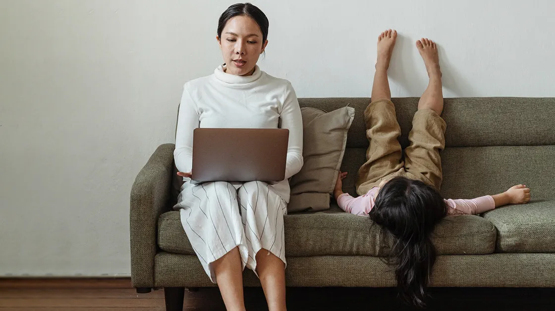 Mother with laptop sitting next to little girl laying on sofa