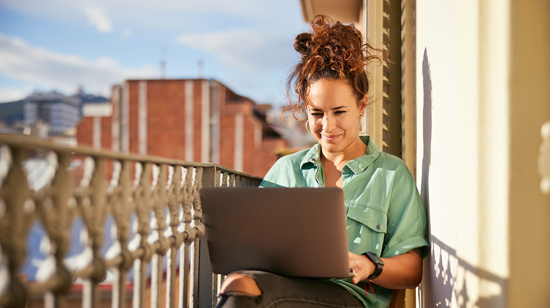 Young woman working on laptop on a sunny balcony. She's wearing a relaxed pale green shirt with short sleeves and her hair is tied up in a big, messy topknot.