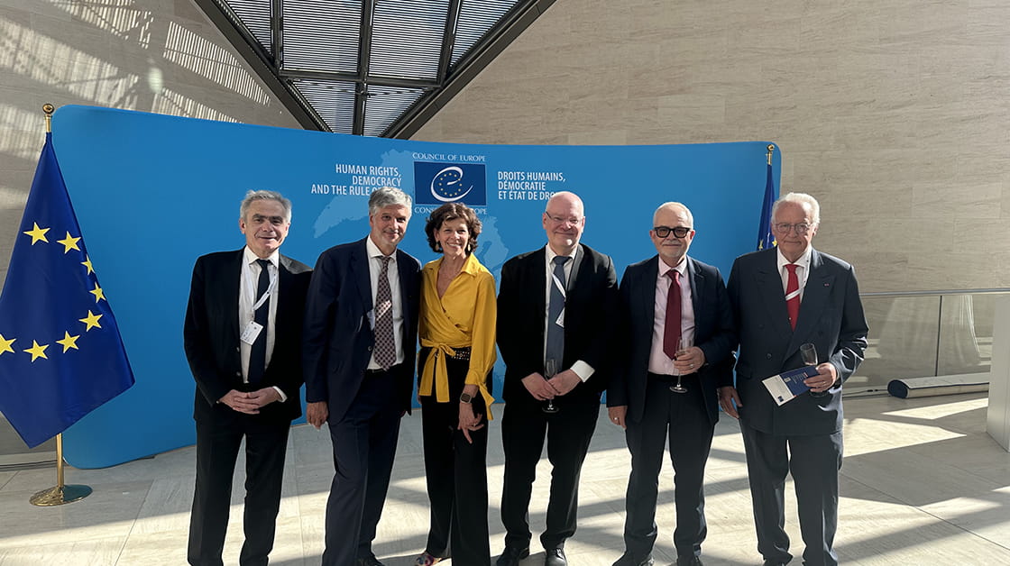 Five white men in suits and one white woman in a yellow blouse and black trousers are standing in front of a blue banner at the Council of Europe. There are two EU flags behind them. They are all smiling. 