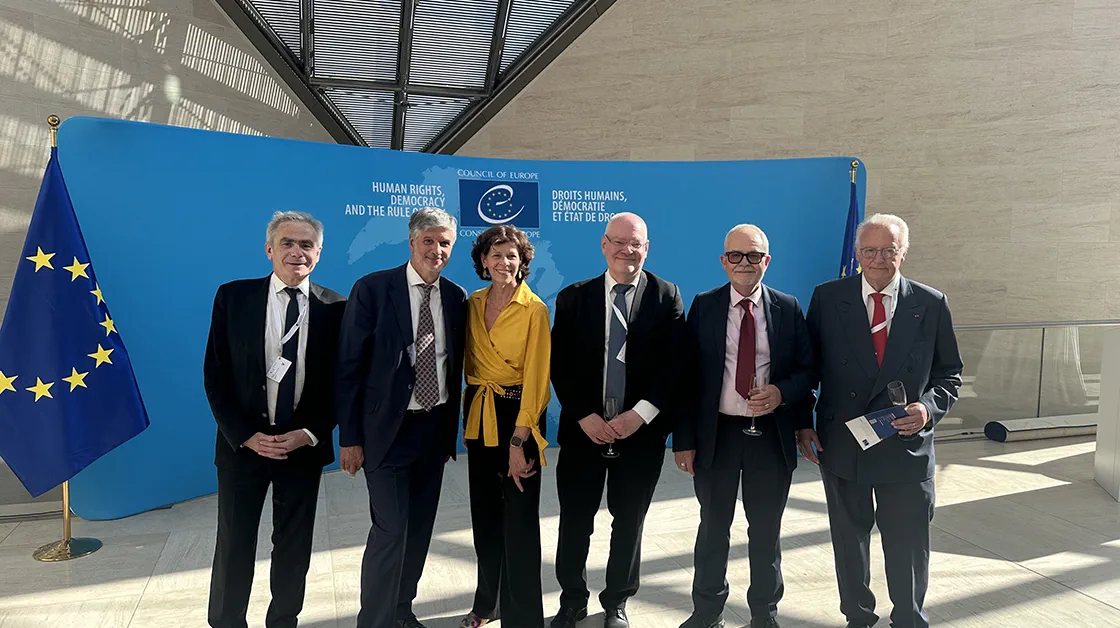 Five white men in suits and one white woman in a yellow blouse and black trousers are standing in front of a blue banner at the Council of Europe. There are two EU flags behind them. They are all smiling.